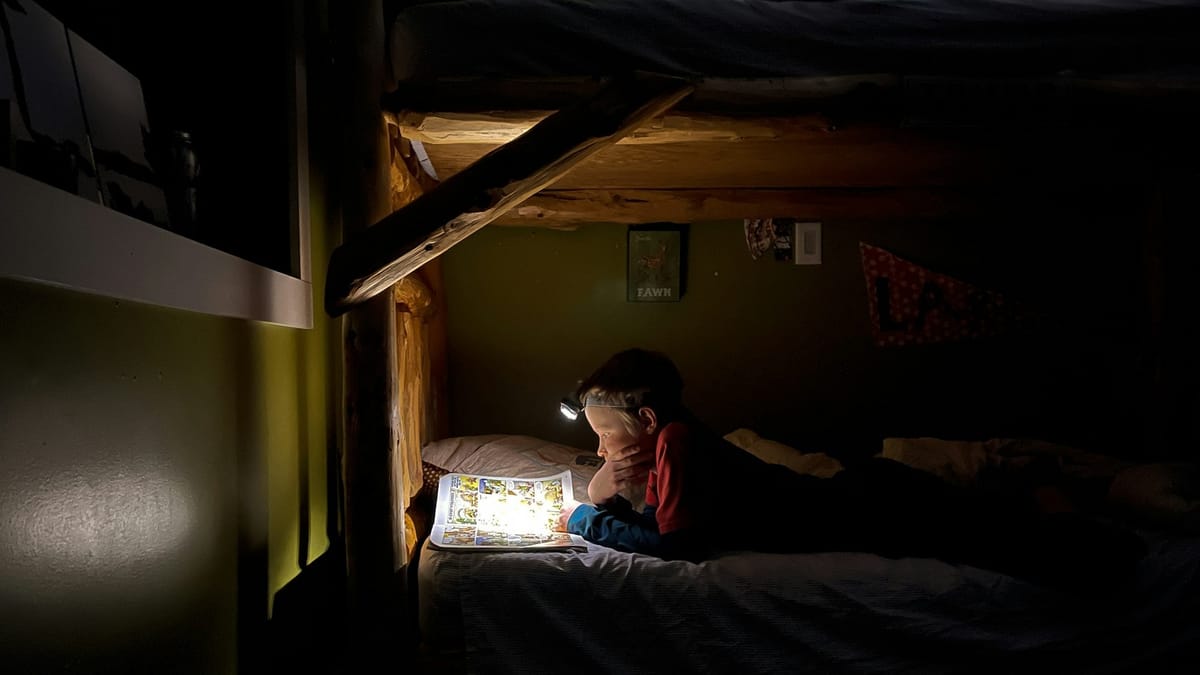 A child reads a comic book in bed with a headlamp.